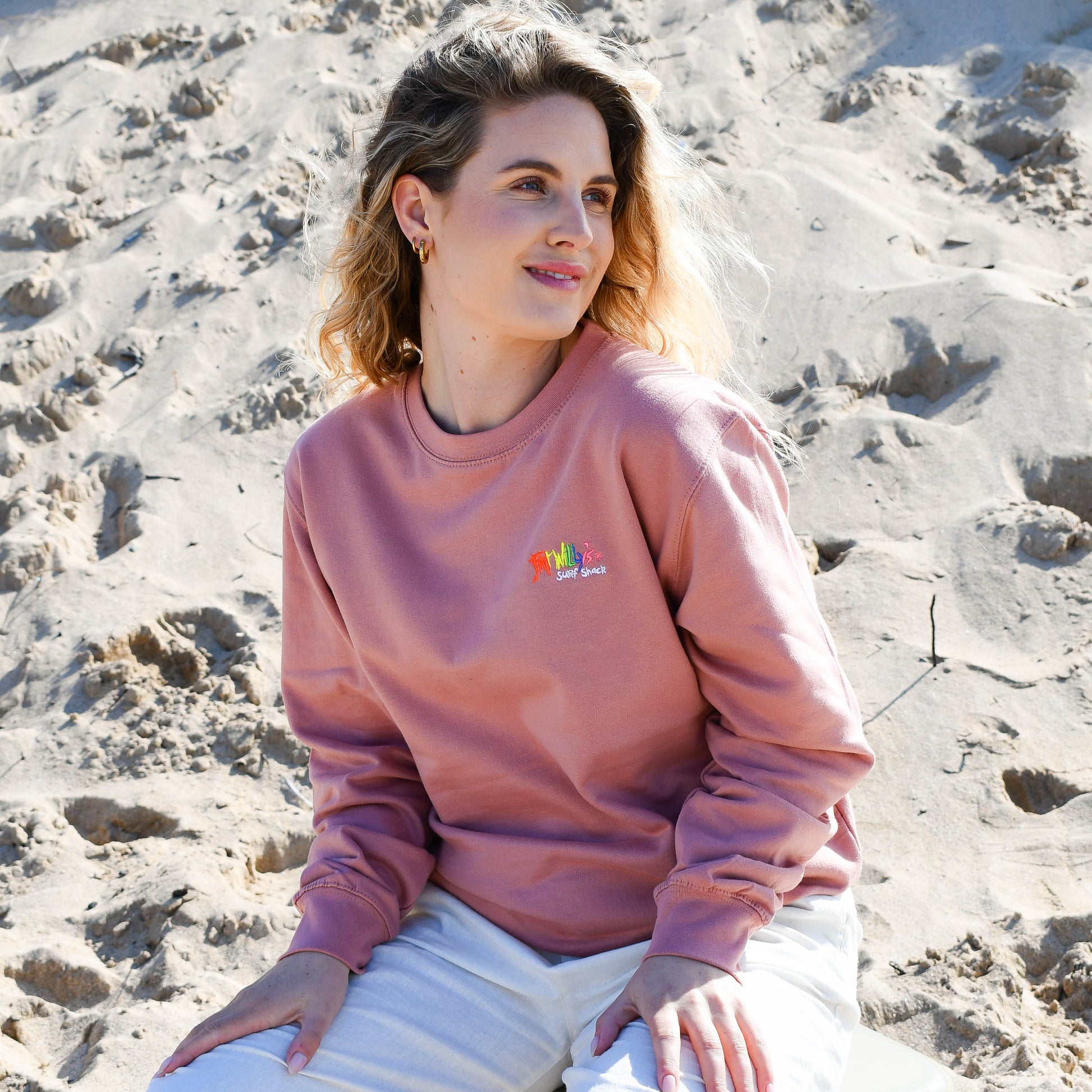 Woman sitting on a beach wearing a pink sweatshirt with a colorful logo.