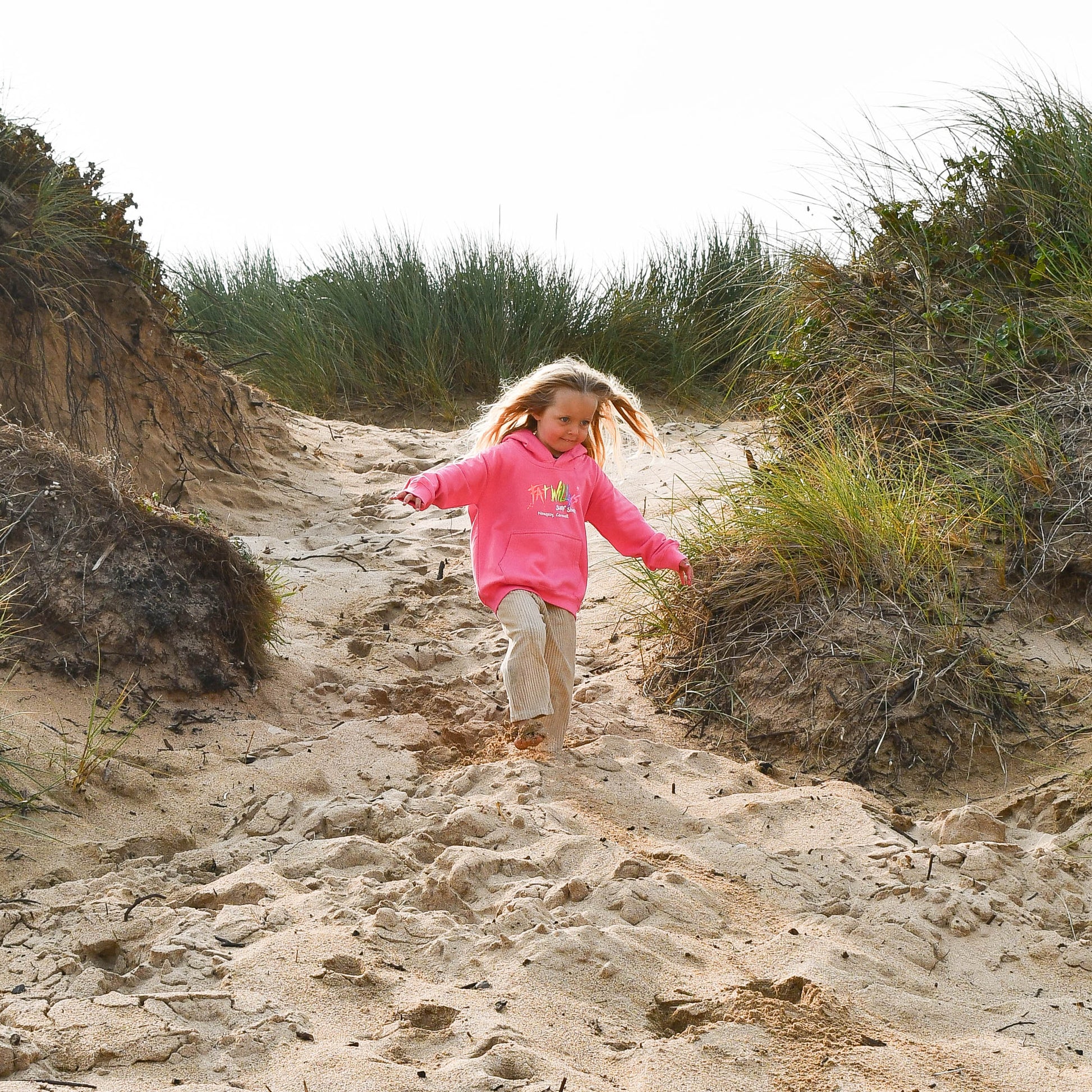 Child in a pink jacket running on a sandy path with grass and shrubs around