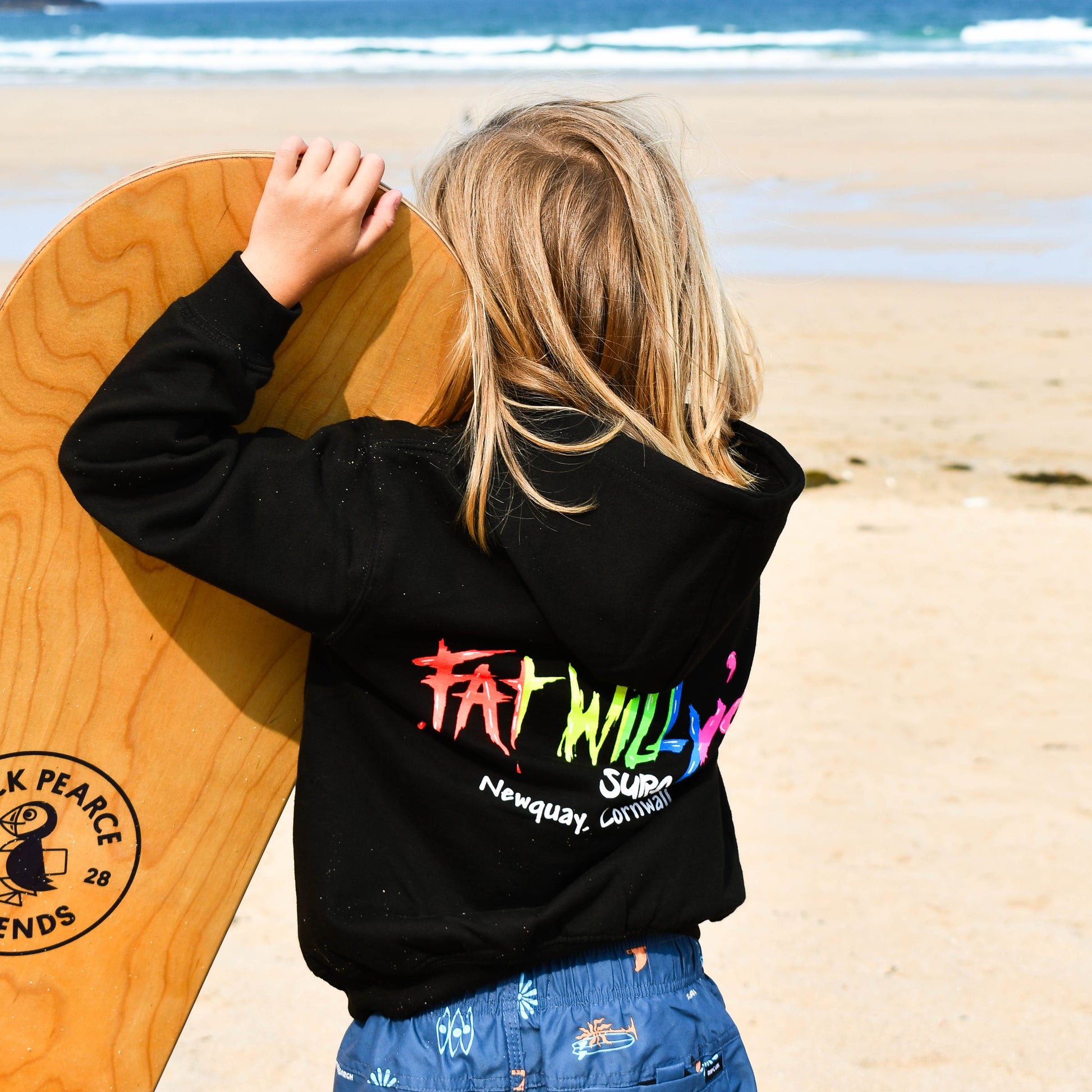 Child holding a wooden surfboard on a beach with a colorful 'Fat Willie' logo on a black sweatshirt.