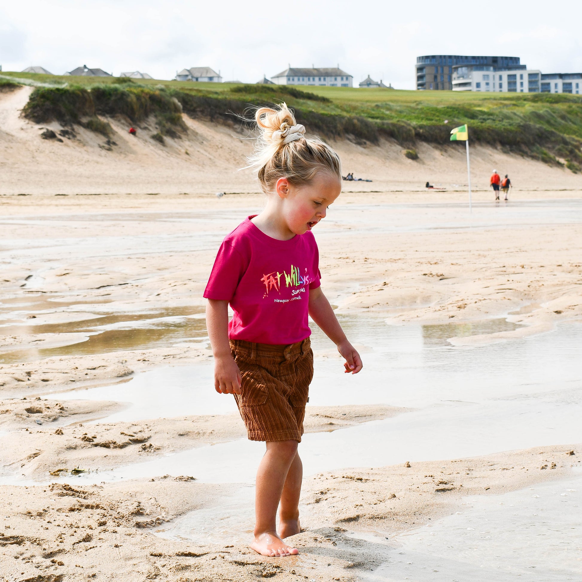 Child in pink shirt and brown shorts standing on a sandy beach with grassy dunes in the background.