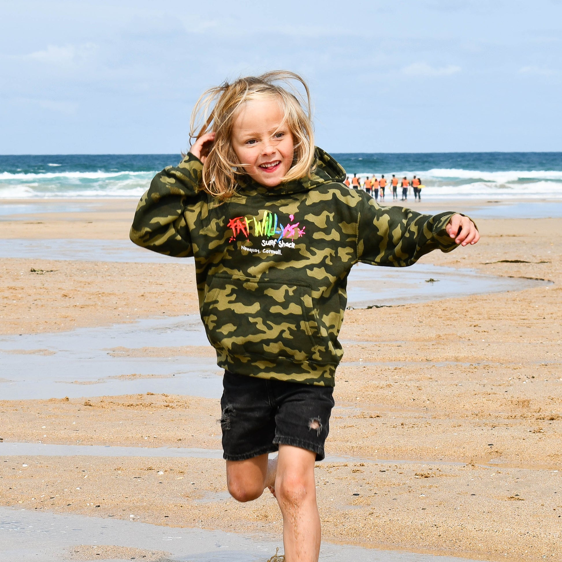 Child running on a beach wearing a camouflage hoodie with colorful text