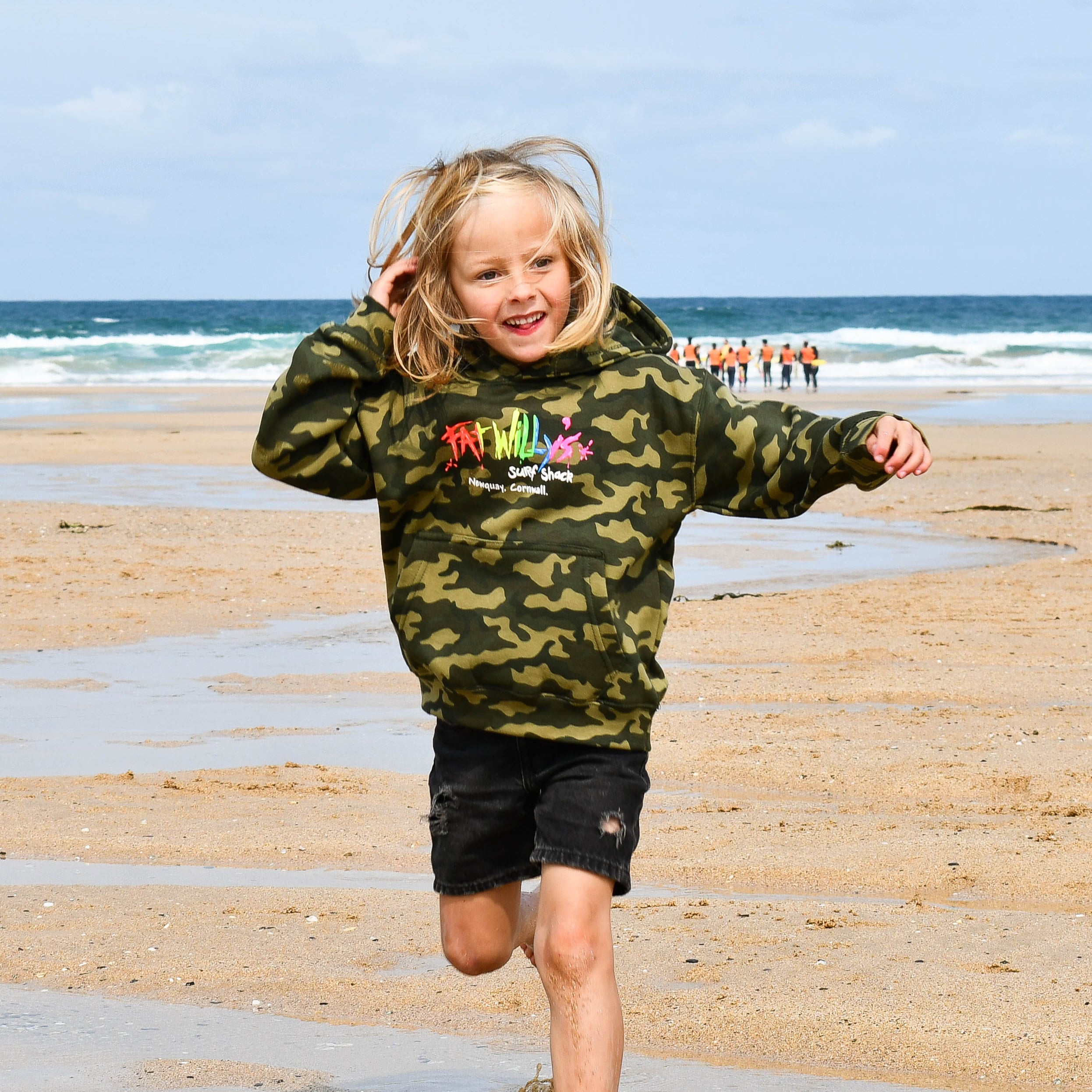 Child running on a beach wearing a camouflage hoodie with colorful text