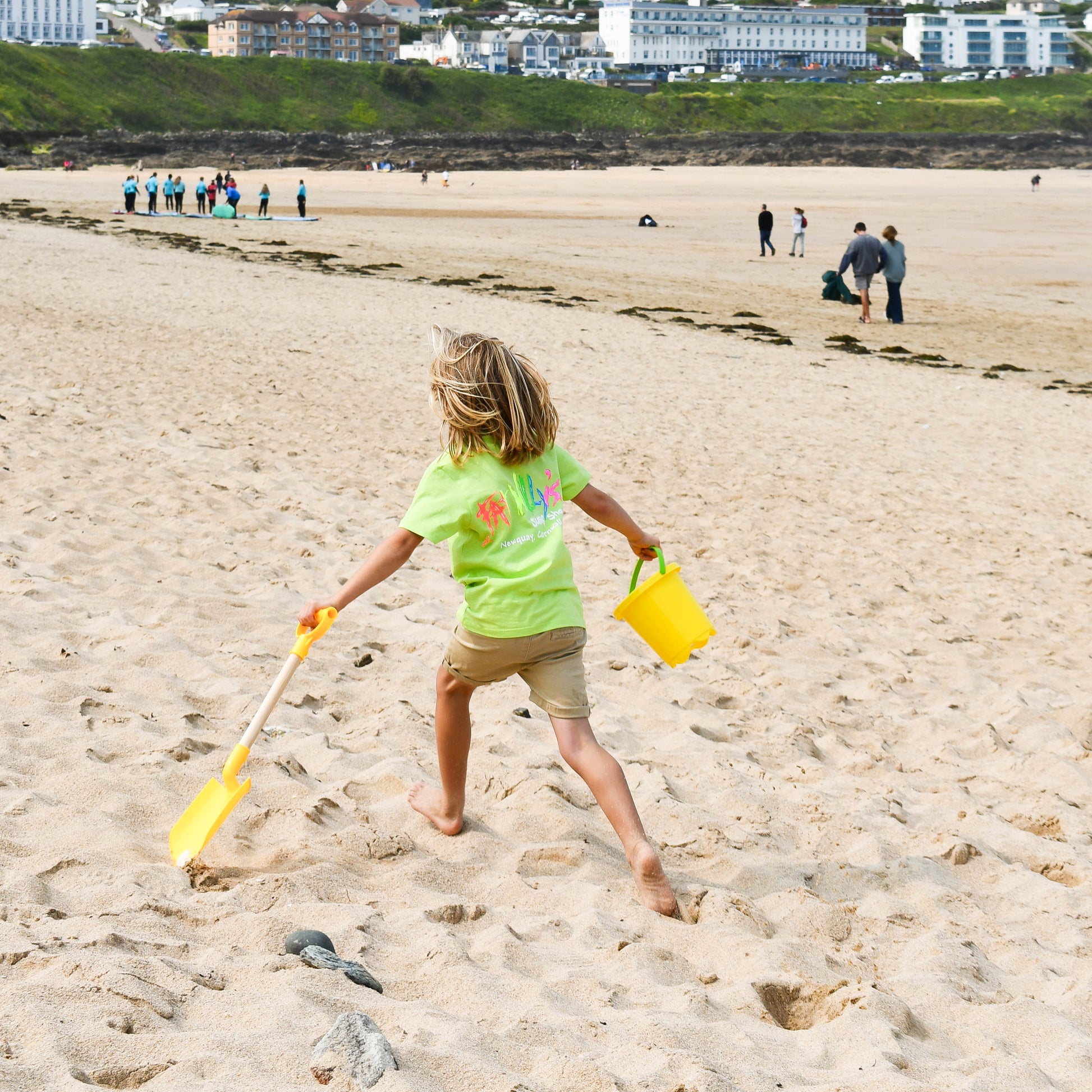 Child playing on a sandy beach with a yellow bucket and spade, buildings in the background.