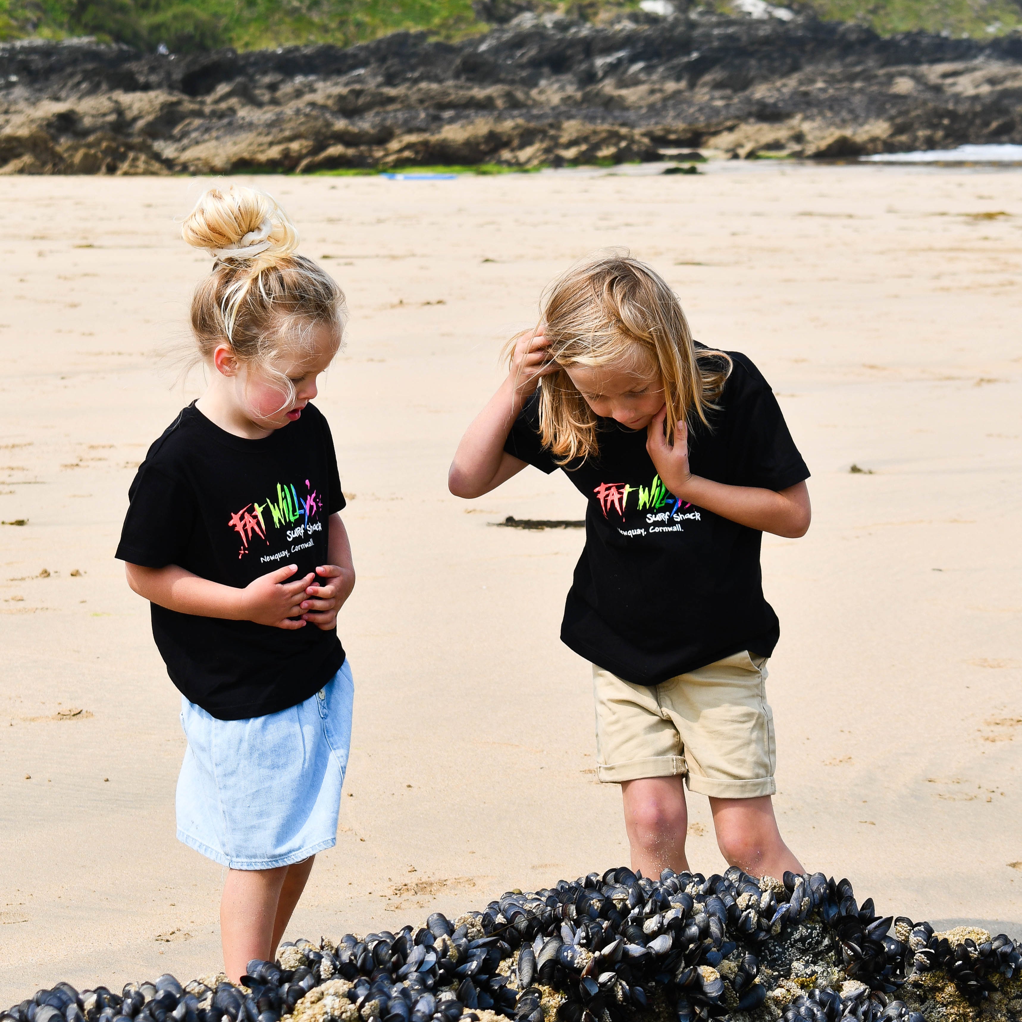 Two children on a beach wearing matching black t-shirts with colorful text.