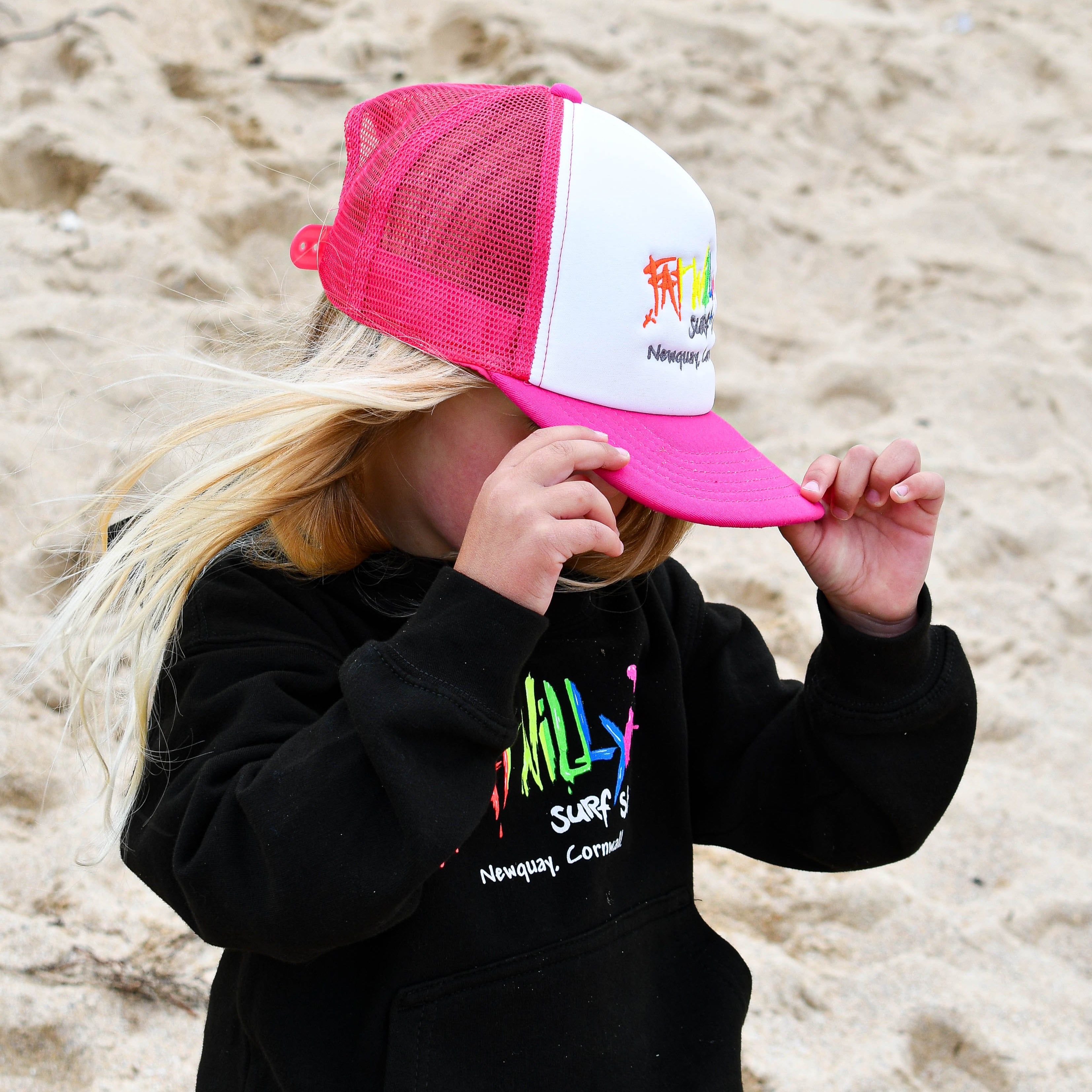 Child wearing a black hoodie with colorful text and a pink and white cap on a sandy background