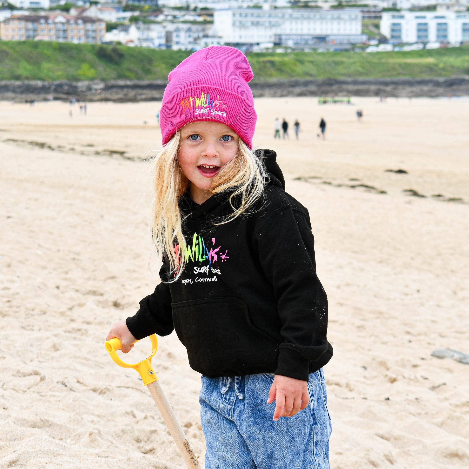 Child wearing a pink beanie and black hoodie on a beach with buildings in the background