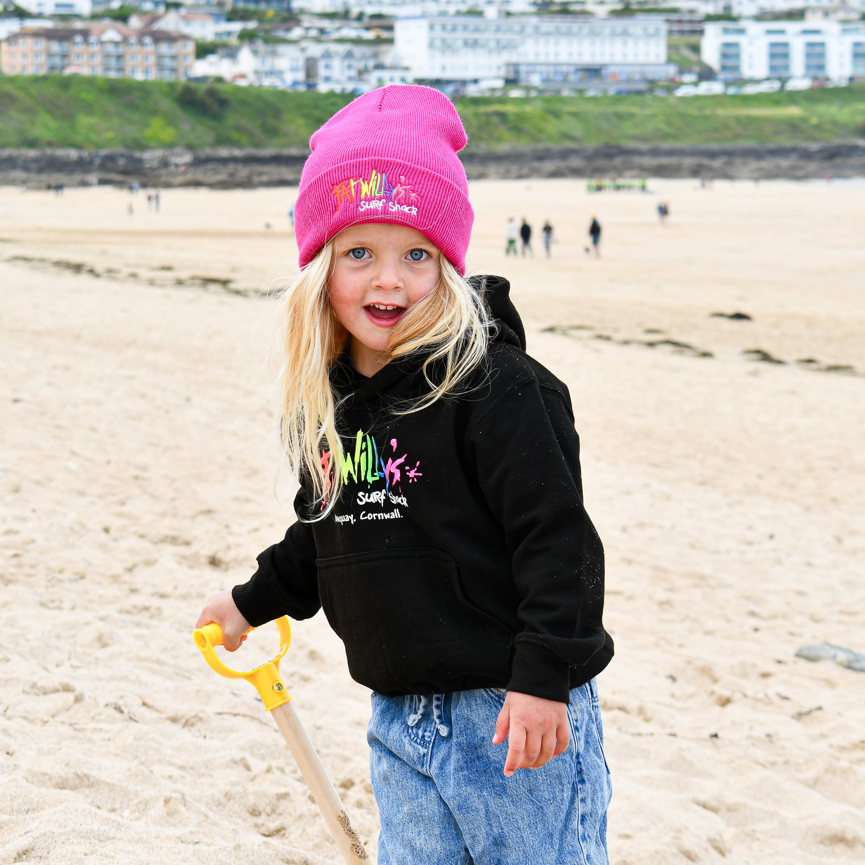 Child wearing a pink beanie and black hoodie on a beach with buildings in the background