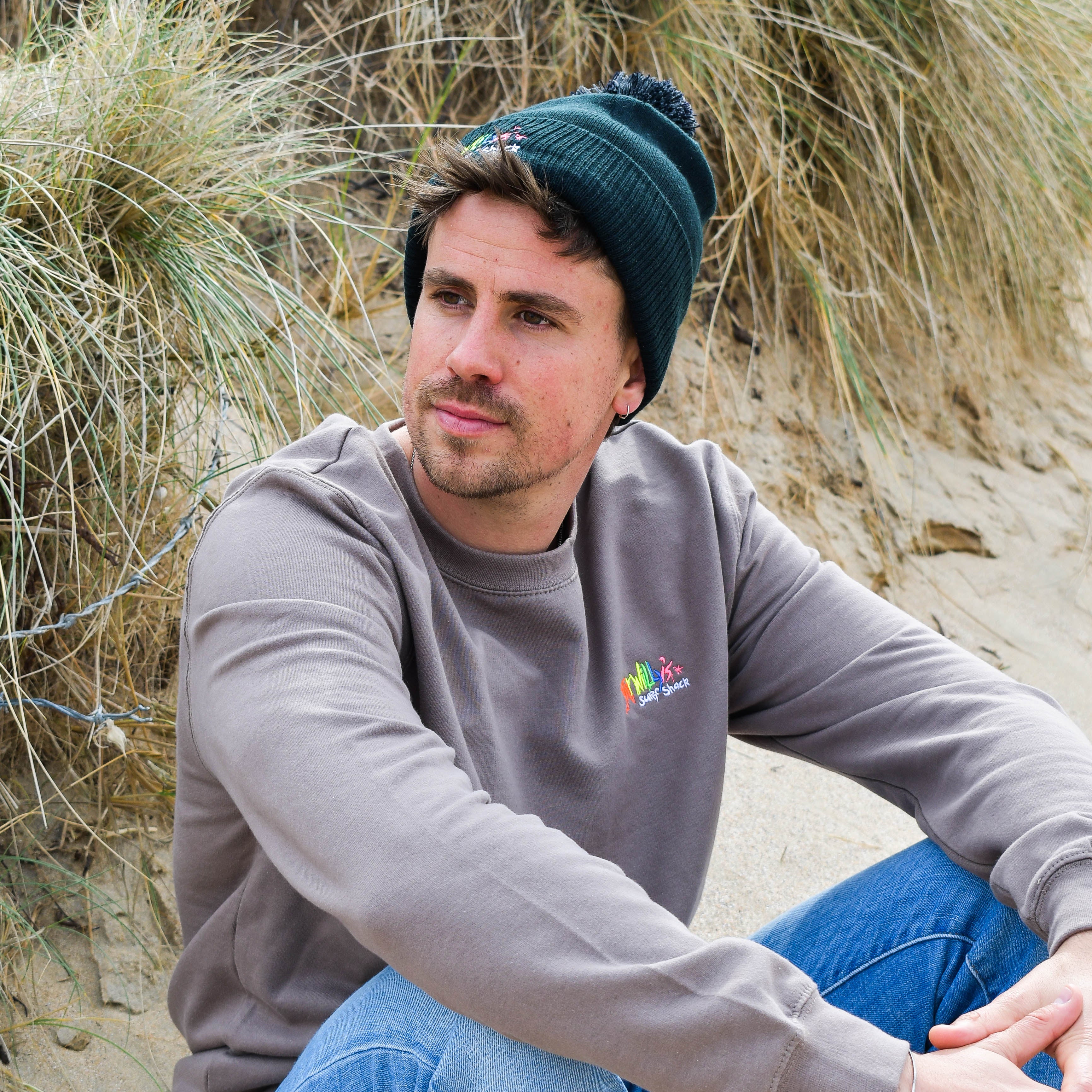 Man sitting on a sandy beach wearing a gray sweatshirt with a logo and blue jeans.