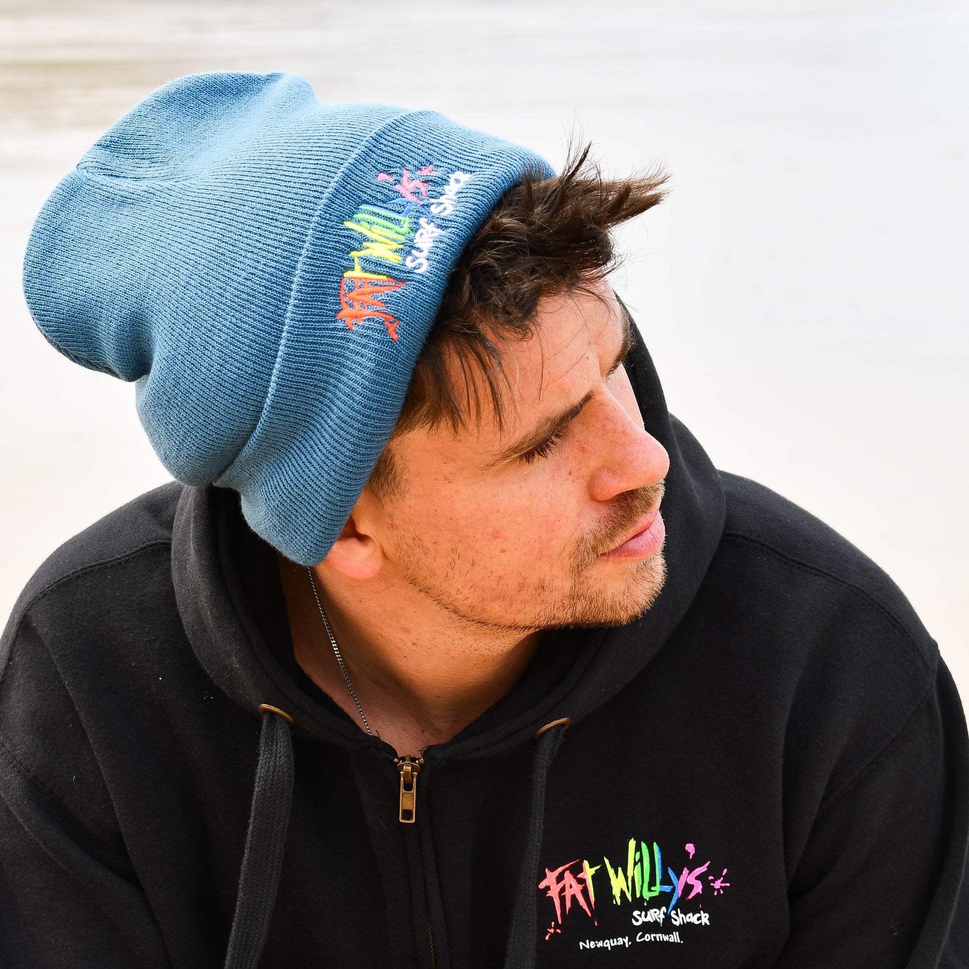 Person wearing a black hoodie with colorful text and a blue beanie with embroidered text, sitting by a body of water.