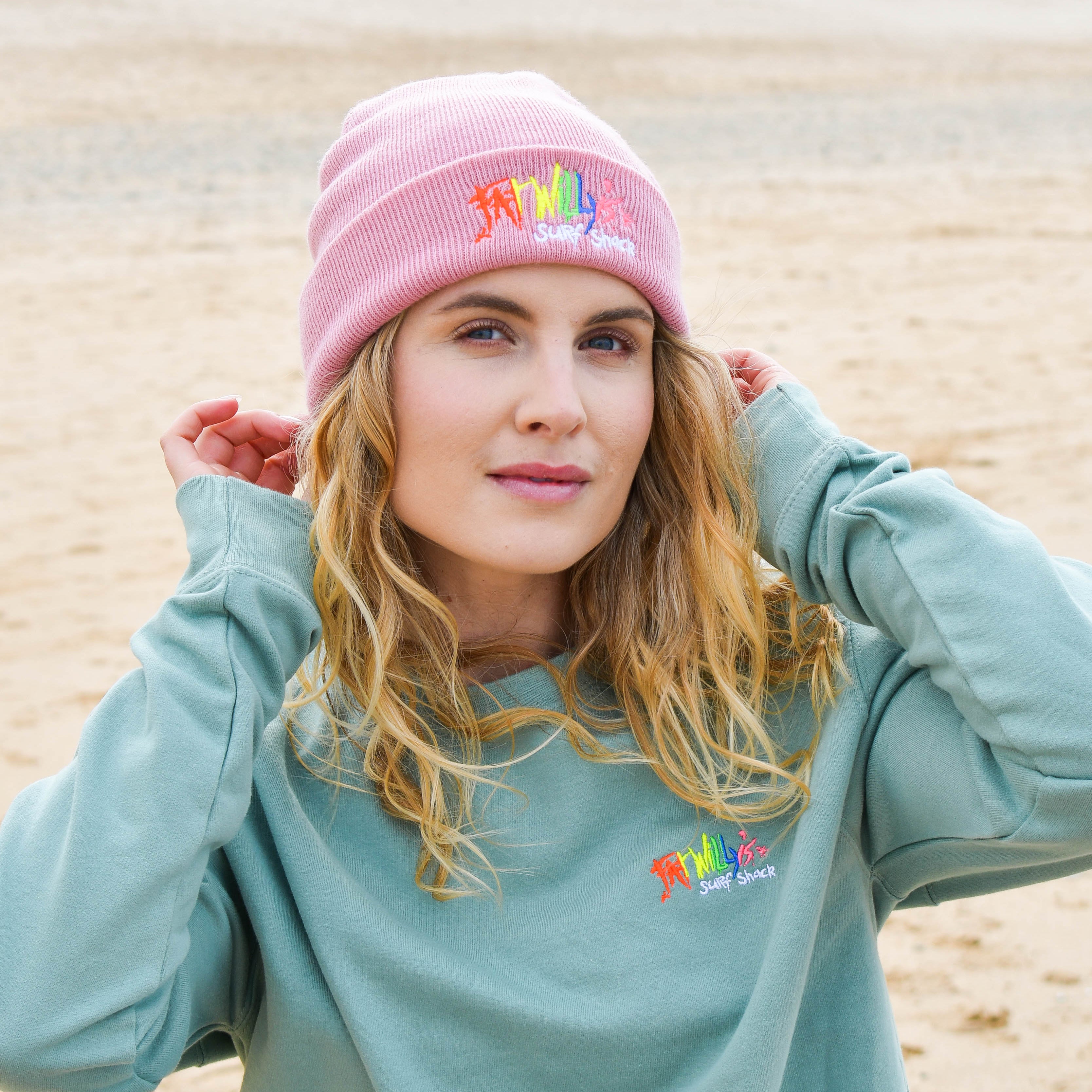 Woman wearing a light blue sweatshirt and pink beanie with colorful text on a beach.
