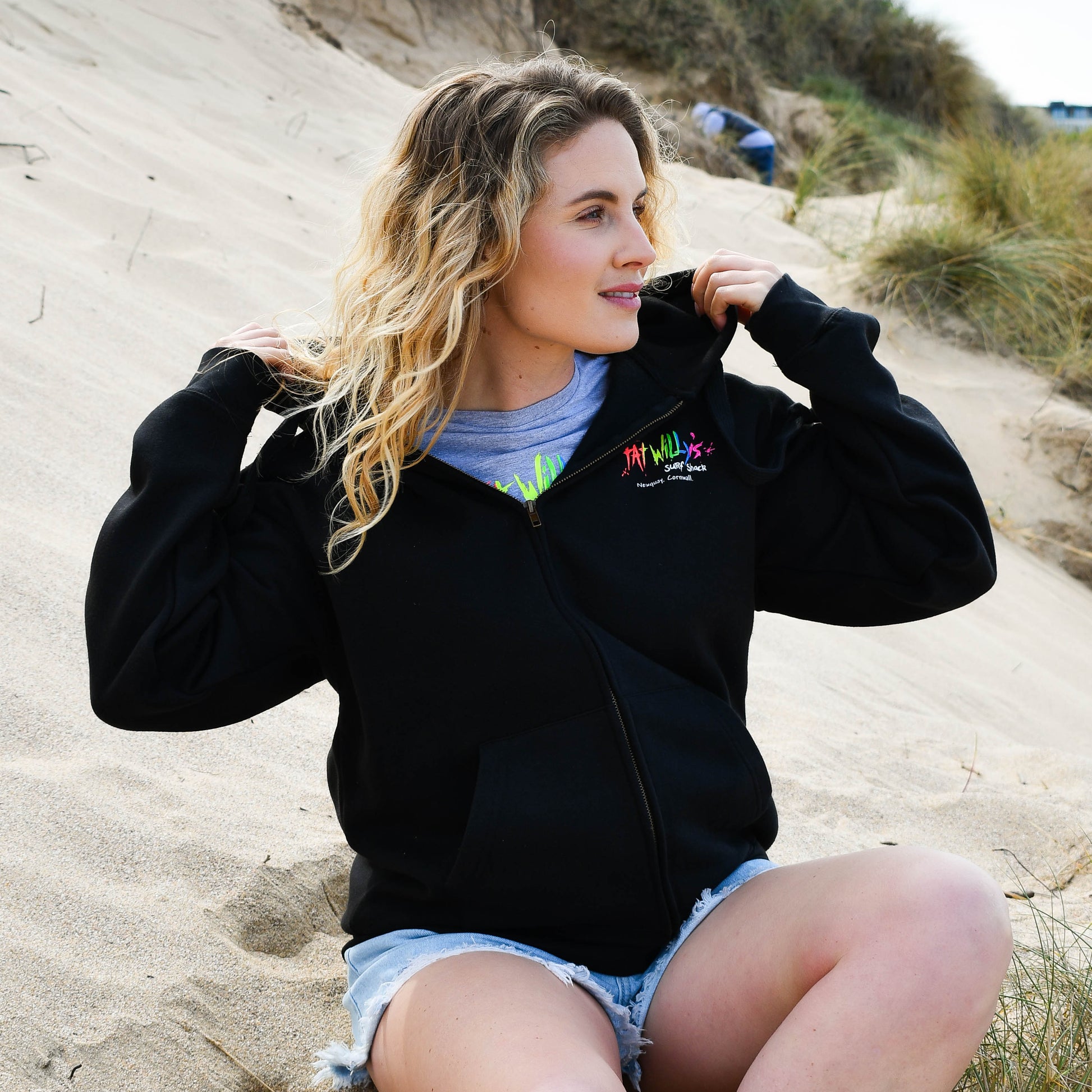 Woman sitting on a sandy beach wearing a black jacket with a logo.
