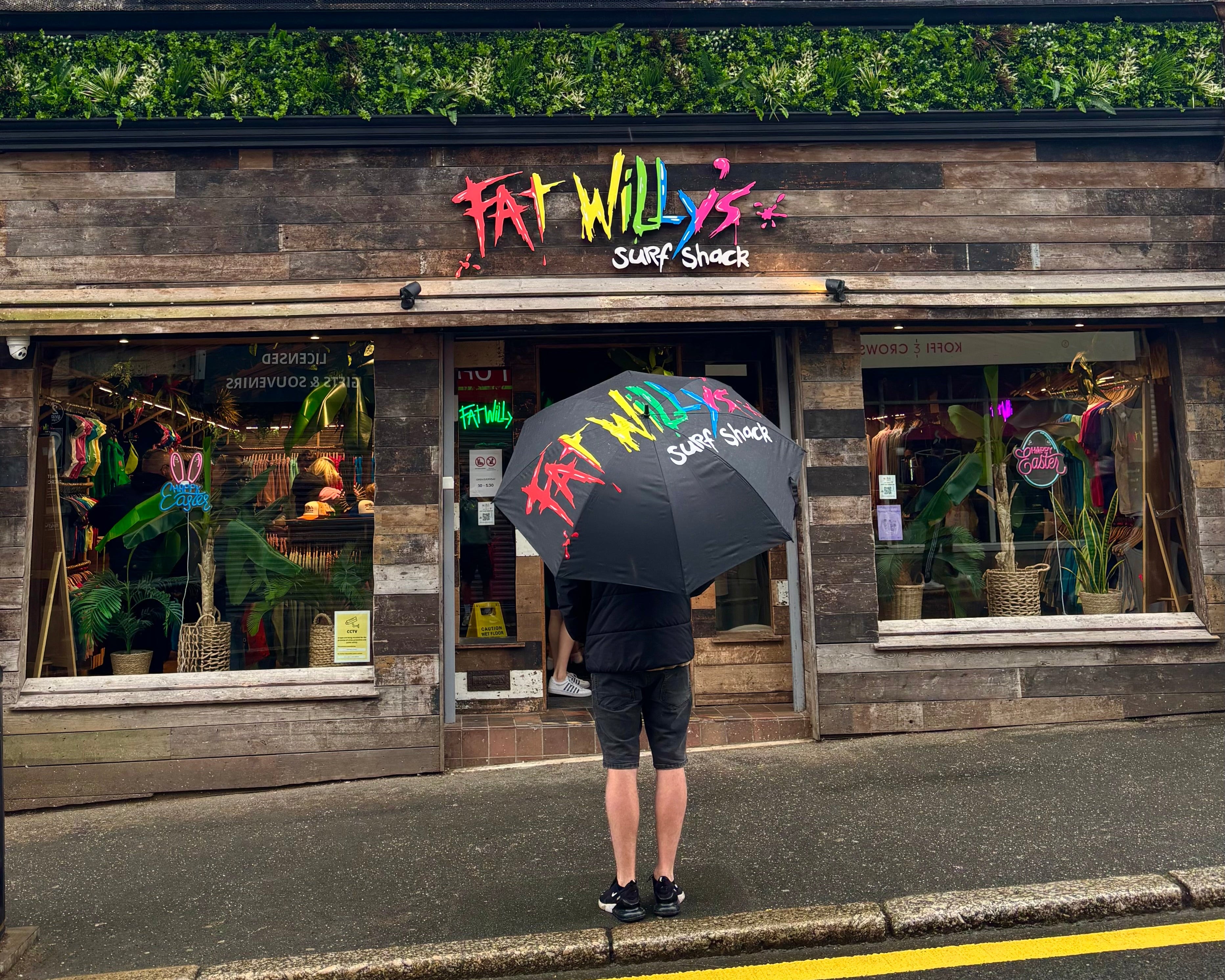 Person with a colorful umbrella standing on a street in front of Fat Willy's Surf Shack.