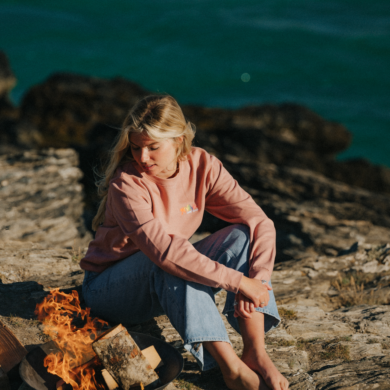 Person sitting by a campfire on a rocky beach with ocean in the background