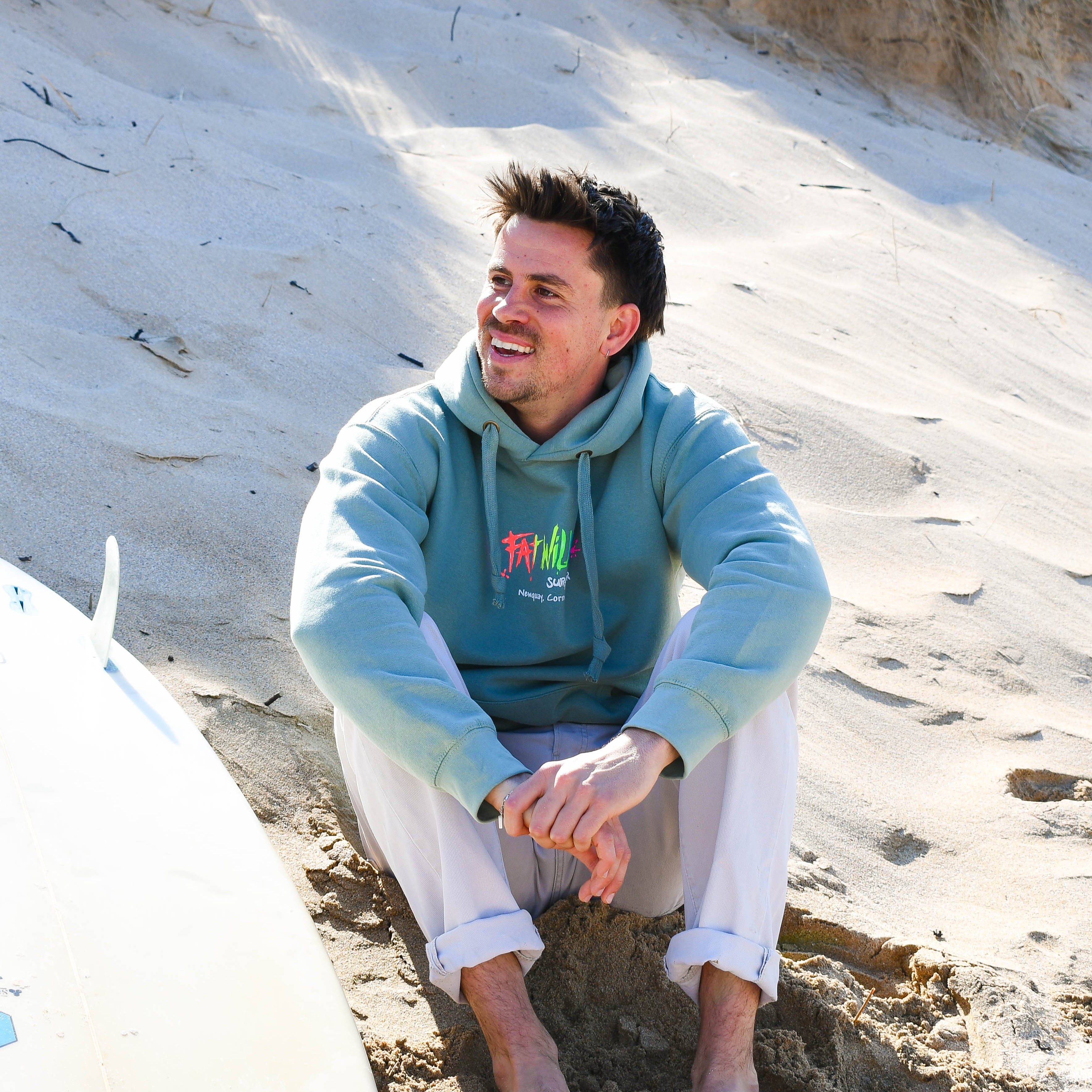 Man sitting on a sandy beach wearing a sage green hoodie with a Fat Willy's logo.